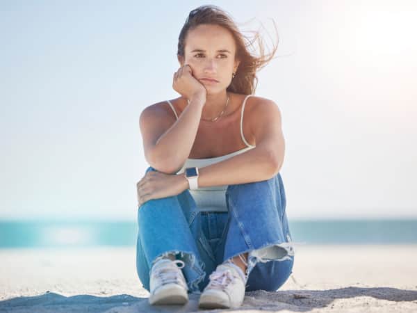 A woman sitting on the beach, ocean in the background, looking towards camera with somber expression